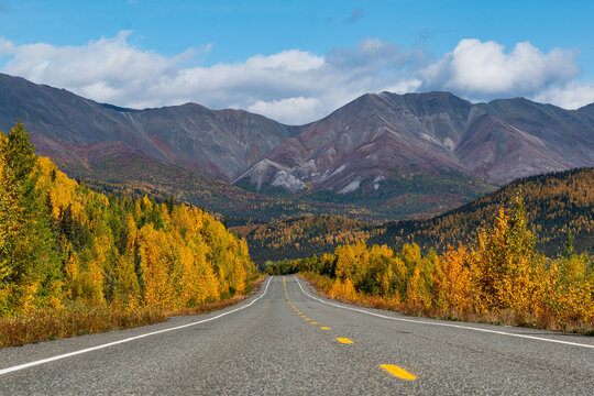 Two lane highway through yellow fall color trees under blue sky with puffy clouds with mountain range in the distance