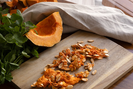 Close up of raw orange Kabocha pumpkin seed on a wooden board. Blurred background with a pumpkin quarter, fresh parsley and beige cloth. Daylight, warm colors, selective focus 