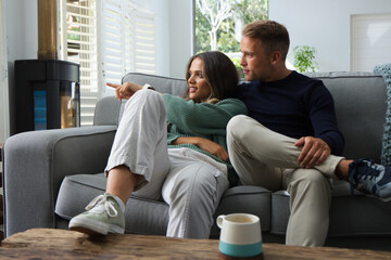 Obraz premium Couple sitting closely on gray sofa in living room, woman pointing left, ceramic mug nearby