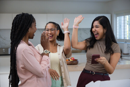 Three female friends high-fiving and laughing at kitchen island with maroon smartphone and watches