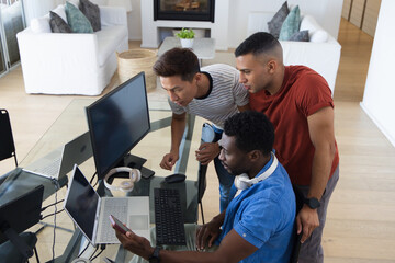 Obraz premium Diverse male coworkers leaning over glass desk in living room workspace, viewing phone and laptops