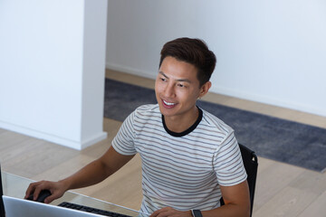 Obraz premium Mid-adult Asian man smiling and working at desk in bright modern room with laptop, mouse
