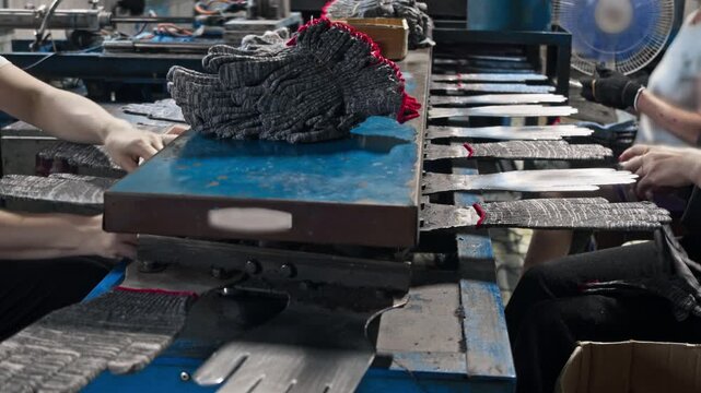 Close up shot reveals textile factory workers placing knitted grey work gloves onto metal shaping molds. Industrial manufacturing line processes protective hand wear during busy production shift.