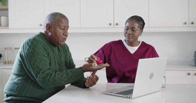 African American caregiver checking laptop, pouring pills to patient taking meds at kitchen island
