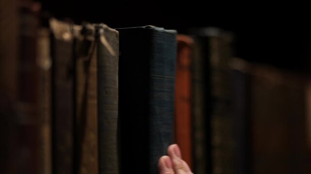 Close-up of a hand selecting an old book from a dimly lit library shelf