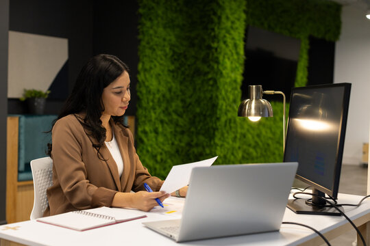 Woman reviewing documents at modern office desk, wearing brown blazer, holding blue pen, laptop