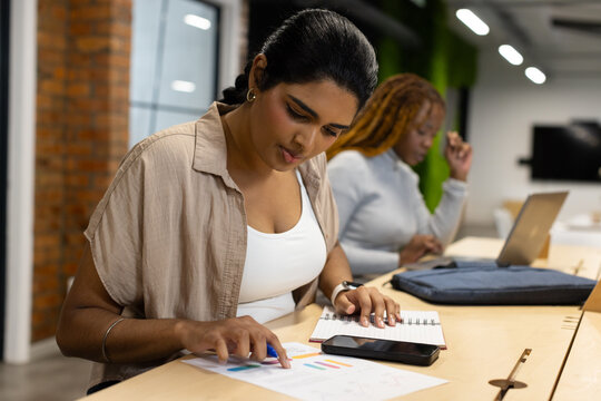 Diverse female coworkers leaning over printed charts and smartphone pointing at graphs in office