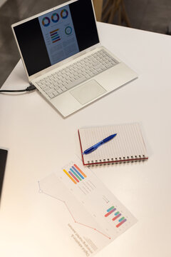 Open laptop showing charts on table, charger trailing, notebook beside printout, backdrop blurring