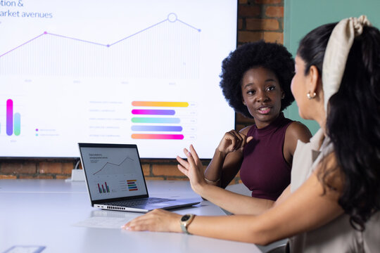 Diverse female coworkers in blazers discussing laptop and wall display charts in conference room