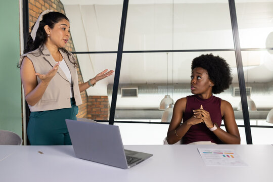 Diverse women staff in office wear presenting and listening at office table with laptop and charts