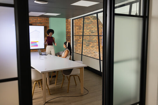 Diverse female coworkers presenting chart and discussing data at white conference table with laptop