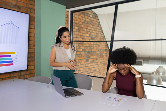 Hispanic and African American women in work attire reviewing chart and laptop at meeting table