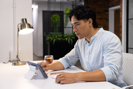 Asian man wearing light-blue shirt sitting at desk in modern office using tablet and brass lamp