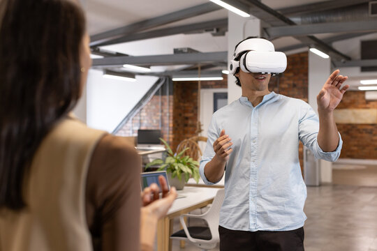 Diverse team Asian man wearing white VR headset gesturing while woman holding phone in open office