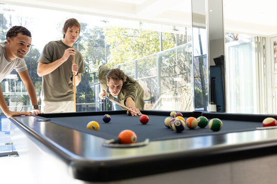 Diverse adult male friends taking shot over pool table with billiard balls by glass doors