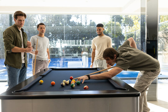 Diverse male friends playing pool in modern game room, holding cues, leaning over navy-felt table