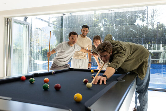 Diverse men lining up shot over black-felt pool table with billiard balls at home holding cues