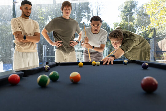 Diverse male friends playing on navy felt pool table in lounge near glass walls with cues