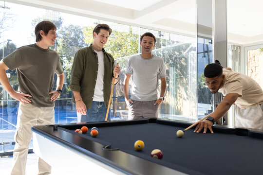 Diverse male friends playing pool in home recreation room around black-felt table, holding cues