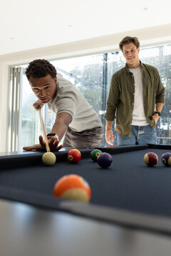 Diverse male friends playing pool, lining up shot on pool table with cue near sliding doors