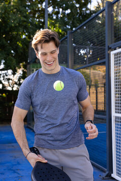 20s man bouncing yellow ball with black paddle on blue court near fence wearing shorts