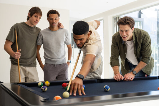 Diverse male friends playing billiards in rec room, shooter aiming cue with scattered balls