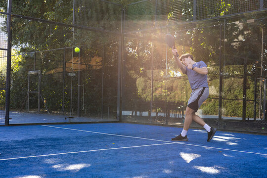 Man in his 20s playing padel at blue court, raising racket toward yellow ball, copy space