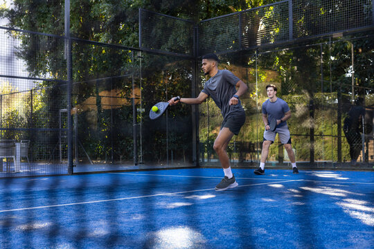 Diverse male partners wearing grey shirts playing padel on blue court with rackets, hitting ball
