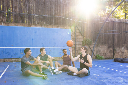 4 adult men in sportswear sitting on blue court spinning basketball and talking near portable hoop