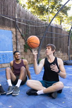 Diverse male friends sitting on blue court with hoop, one spinning basketball on index finger