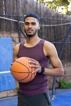 Adult male holding orange basketball with smartwatch on left wrist in maroon tank on blue court
