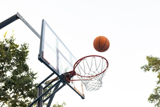 Orange basketball is descending toward red rim on outdoor hoop, leafy trees flanking