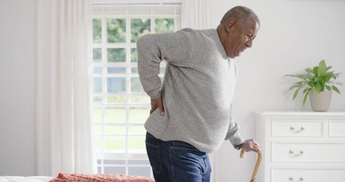 Senior African American man pressing lower back, standing from bed and walking with cane in bedroom
