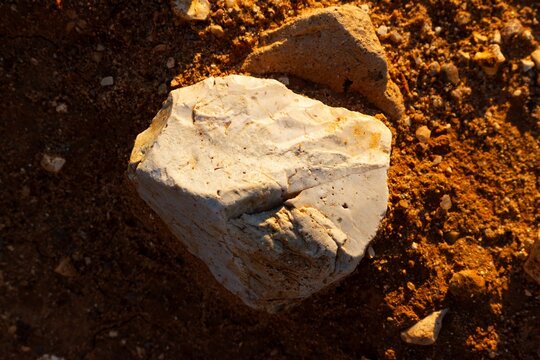A sharp white flint stone lying on dry golden sand inside a sunny quarry, macro texture of stone with natural weathered edges under bright warm light during golden hour