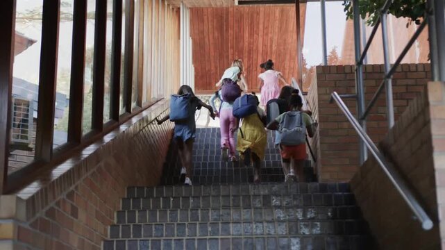 Diverse group of schoolgirls running up covered outdoor staircase, backpacks visible from rear low angle in bright daylight with eager smiles and shared momentum together