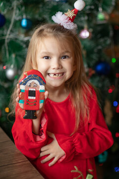 Cute funny little girl with missing primary milk teeth smiling cheerfully and holding a Nutcracker gingerbread cookie near a decorated Christmas tree. Cute child in a red festive dress