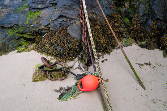 Ropes, rusty chains and a bright orange buoy used for boat berthing sit on the sandy beach against dark rocks in Portsoy, Moray, Scotland