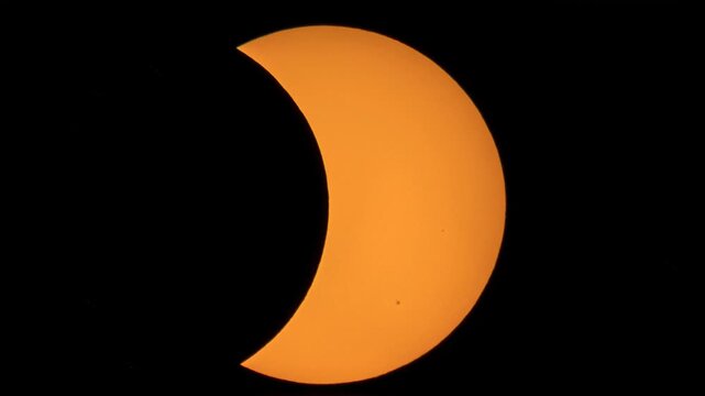 Close-up of a partial solar eclipse, with the dark silhouette of the moon obscuring the left side of the orange sun, highlighting visible sunspots