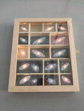 Wooden shadow box displaying miniature painted spiral galaxies inside compartments