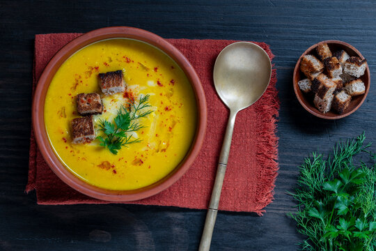 Top view yellow pumpkin cream soup with croutons. Vegetarian lentil or squash puree soup garnished with croutons paprika and fresh herbs in clay bowl on red napkin cozy winter comfort food backdrop