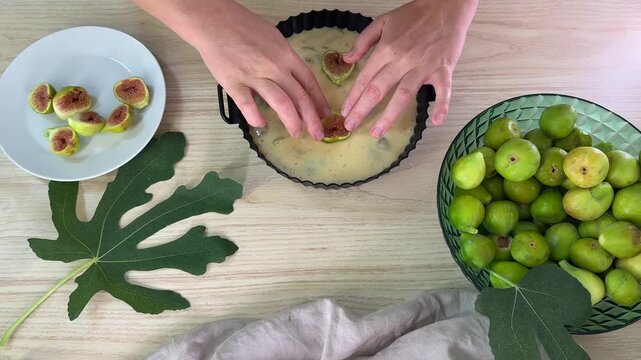 A person places sliced figs into a fluted cake pan filled with batter, surrounded by whole green figs in a bowl and sliced figs on a white plate