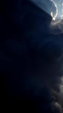 Towering billowing clouds drift across bright blue sky as darker formations move in front of sunlight, revealing glowing silver edges, deep shadows, and slanting sun rays
