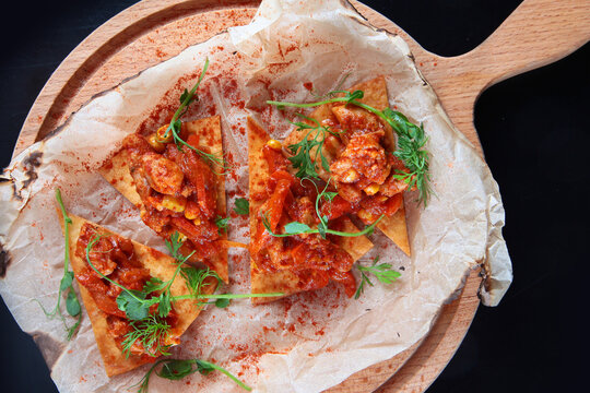 Mexican nachos appetizer with tomato sauce and herbs. Triangular nacho chips with tomato sauce, corn, and herbs on parchment paper. Dish is served on wooden board. Top view