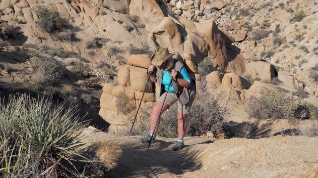 Senior woman wearing a hat and using trekking poles hiking uphill on winding dirt path through a rocky desert canyon, showcasing an active and healthy outdoor lifestyle during retirement