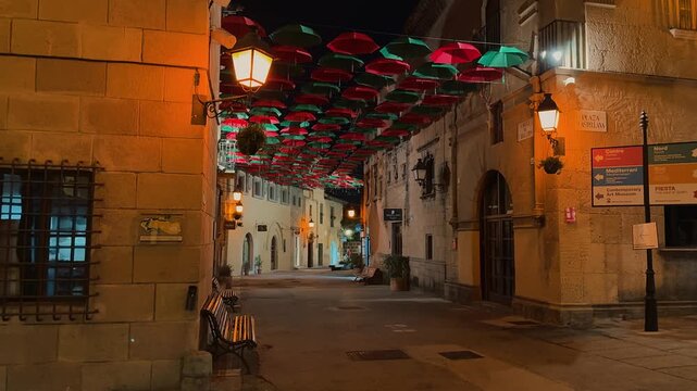 Explore the illuminated streets of Poble Espanyol at night in Barcelona, showcasing traditional Spanish architecture, fortified medieval gate, decorative hanging umbrellas, historic facades,