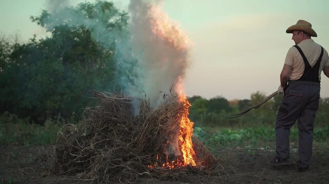 Caucasian man in straw hat and overalls tending to a burning pile of brush, using a pitchfork to manage flames and smoke in a rural outdoor setting during sunset