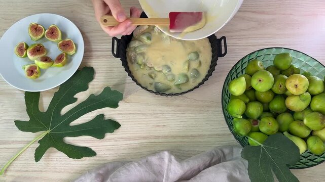 A person pours pale yellow batter from a white bowl into a black fluted cake pan, surrounded by sliced figs on a plate and whole green figs in a bowl