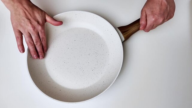 Two pairs of hands interact with a round light colored frying pan resting on a smooth white surface; the hands are holding the handles of the cookware in a repeating sequence