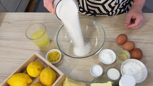 A person wearing a patterned black and white shirt pours white granulated sugar from a glass into a large clear mixing bowl on a light wooden table