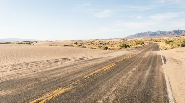 Desert highway disappears into sand dunes with fading yellow lines, suggesting journey and isolation under a clear sky